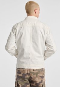 White denim jacket with a tailored fit, visible stitching, and a notched collar. Worn over camouflaged cargo pants. Back view.