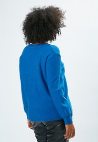 Blauer Pullover mit einer strukturierten Oberfläche und subtilen Schimmereffekten, Rundhalsausschnitt und gerippten Bündchen. Getragen mit schwarzen Hosen.