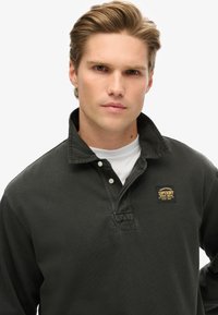Black long-sleeve polo shirt with a textured collar, a white inner shirt, and a small yellow and black brand logo on the chest.