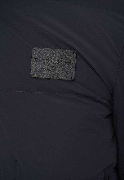 Zilā neilona jaka ar ādas etiķeti, uz kuras rakstīts "Emporio Armani Milano." Gluda tekstūra ar minimālu šuvju detaļu skaitu.