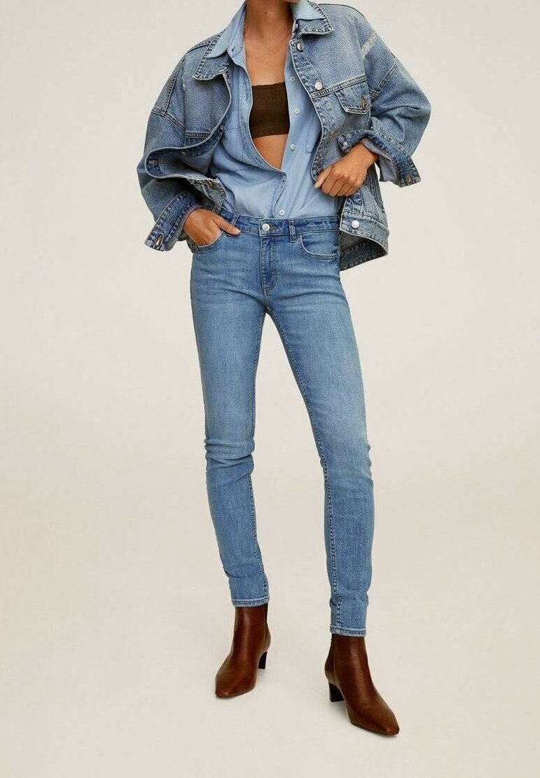 Chaqueta de mezclilla azul claro sobre una camisa azul clara, combinada con unos vaqueros ajustados azul claro y botas de tobillo de tacón marrones. Tejidos texturizados en toda la atuendo.