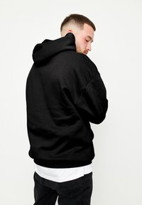Felpa nera con una vestibilità rilassata, tessuto texture, cappuccio grande e polsini a coste, indossata sopra una t-shirt bianca e pantaloni neri.
