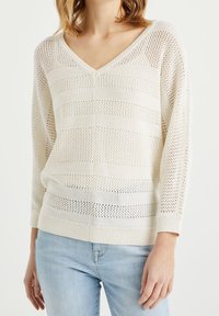 Kevyt neulepusero kermaisen värisenä, jossa on V-pääntie, pitkät hihat ja raidallinen kuvio, joka koostuu avoimista ja kiinteistä segmentistä.