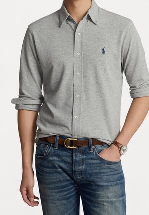 Homme portant une chemise gris clair à boutons avec manches retroussées, jean bleu, ceinture marron, montre en cuir, et une main dans la poche.