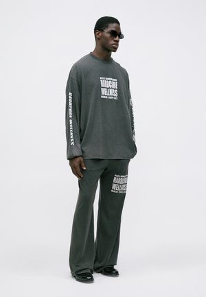 Homem a usar uma camisola larga cinza escuro e calças com texto branco "Hardcore Wellness", sapatos pretos e óculos de sol pretos estreitos, em pé.
