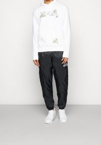 Vit hoodie med gröna grafiska mönster och en framficka, matchad med svarta, glänsande, elastiska byxor som slutar vid anklarna och har en logo. Vita sneakers ingår.