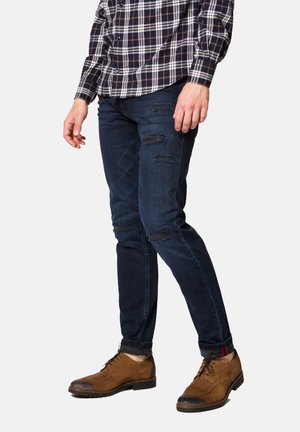 Granatowe przetarte dżinsy o dopasowanym kroju, z wieloma przetartymi miejscami. Zestawione z brązowymi zamszowymi butami i koszulą w kratę.