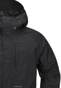 Chaqueta impermeable negra con un cuello alto, bolsillos con cremallera y capucha. Presenta un logo en la parte izquierda del pecho. Textura suave.