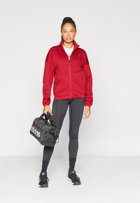 Červená bunda na zips s bočnými vreckami, čierne legíny a čierna športová taška. Bunda má vysoký golier a textúrovanú tkaninu.