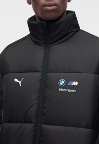 Homme portant une doudoune noire avec les logos BMW Motorsport et Puma sur la poitrine, zippée jusqu'au col
