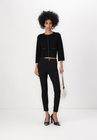 Suéter negro recortado con mangas tres cuartos y detalles de tachuelas metálicas. Pantalones ajustados negros y zapatos de tacón negro brillantes. Bolso de color claro.