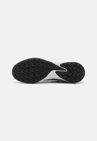 Suela de goma negra con un patrón de dibujo en zigzag profundo en una zapatilla deportiva blanca, mostrada desde la parte inferior sobre un fondo blanco.