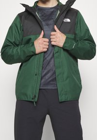 Veste déperlante avec un corps vert et des épaules noires, équipée d'une capuche, d'une fermeture éclair à l'avant et du logo The North Face sur la poitrine.