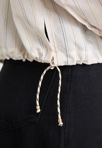 Tessuto a strisce di colore chiaro con orlo elastico arricciato e cordoncino a contrasto con dettagli dorati e finitura testurizzata. Vita scura visibile.
