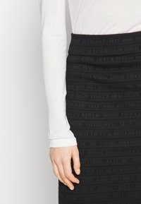 Zwarte aansluitende rok met tekstuurlijke horizontale strepen en reliëftekst. Gekoord over een witte longsleeve top, waarbij een deel van de hand zichtbaar is.