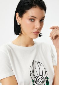 Valkoinen puuvillapaita, jossa on graafinen printti kasvista mustilla viivoilla ja vihreillä yksityiskohdilla. Mallilla on hopeakorvakorut ja lyhyet mustat hiukset.