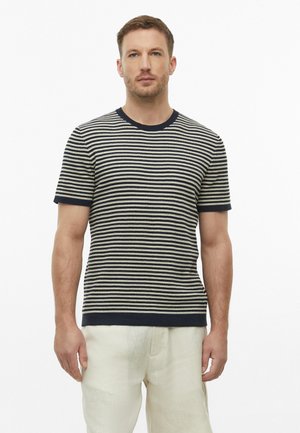 Man with short hair wearing a black and white striped short-sleeve shirt and light beige pants standing against a white background.