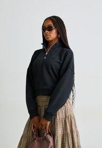 Sudadera negra recortada con cuello con cremallera, combinada con una falda a cuadros. La modelo lleva gafas de sol oscuras y sostiene un bolso rosa.