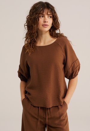 Femme aux cheveux bruns bouclés portant un haut ample marron texturé à manches courtes et un pantalon assorti, mains dans les poches, regardant de côté sur un fond beige.