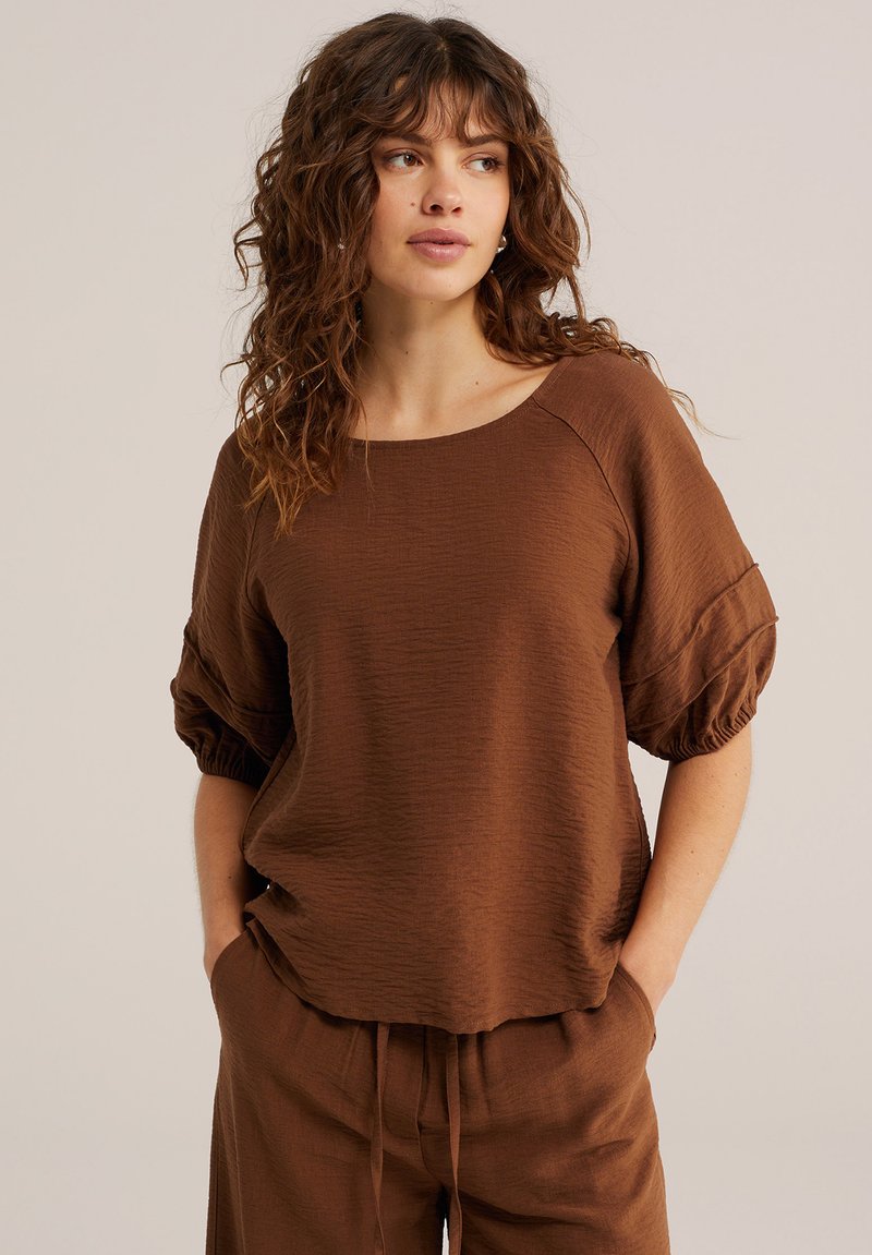 Femme aux cheveux bruns bouclés portant un haut ample marron texturé à manches courtes et un pantalon assorti, mains dans les poches, regardant de côté sur un fond beige.
