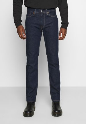 Homme portant un jean bleu foncé droit, un haut noir à manches longues et des bottes en cuir noires, debout sur un sol gris uni.