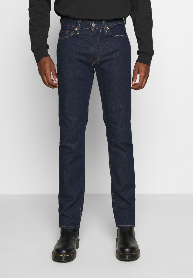 Homme portant un jean bleu foncé droit, un haut noir à manches longues et des bottes en cuir noires, debout sur un sol gris uni.