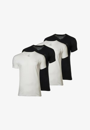 Jeune homme debout pieds nus portant une chemise noire à manches courtes et un pantalon long gris clair sur un fond blanc uni.