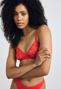 Rode kanten bralette met een driehoekige vorm, dunne verstelbare bandjes en een klein logo-detail. Geplaatst tegen een neutrale achtergrond.