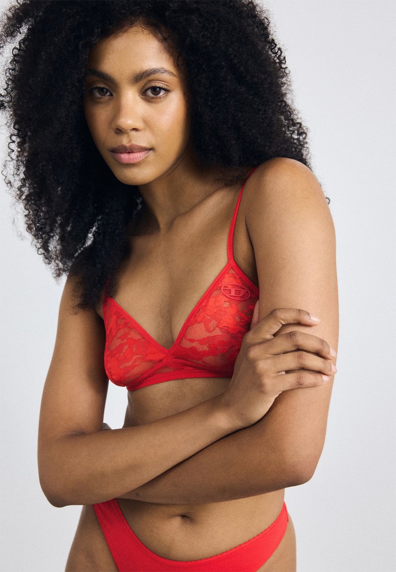 Rode kanten bralette met een driehoekige vorm, dunne verstelbare bandjes en een klein logo-detail. Geplaatst tegen een neutrale achtergrond.