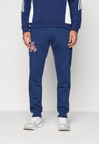 Temno modri športni hlače iz mehkega blaga, z vezenim črko "A" v burgundski barvi na levem stegnu in elastičnimi manšetami na gležnjih.