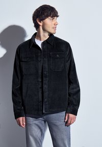 Černá kordurová košile s kapsami na hrudi, zapínání na knoflíky a texturovaná látka, zkombinovaná se světle modrými denimovými džíny.