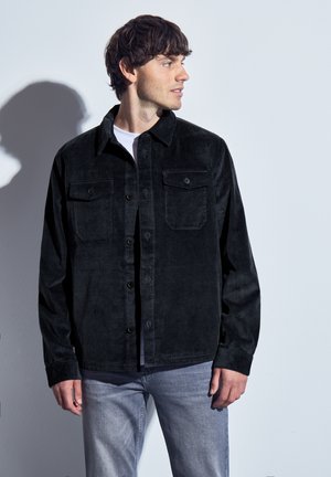 Zwarte corduroy overhemdjack met twee borstzakken, knoopsluiting aan de voorkant en een gestructureerde stof, gecombineerd met lichte denim jeans.