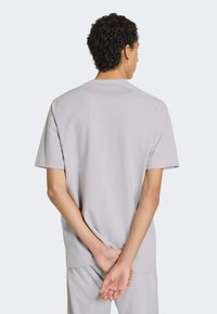 Jasnoszara koszulka z krótkim rękawem o luźnym kroju, gładkiej teksturze i okrągłym dekoltem, widziana od tyłu z założonymi skrzyżowanymi rękami.