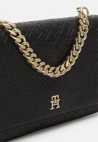 Schwarze Handtasche mit strukturiertem Material, goldfarbener Kettenriemen und graviertem Logo-Detail auf der Vorderseite. Das Material ist mit einem geometrischen Muster geprägt.