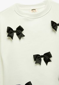 Sudadera blanca con lazos de cinta negra esparcidos en el frente. Textura suave, cuello y puños acanalados, diseño casual para niños.