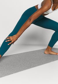 Teal leggings i stretchtyg, högmidjad design med en mjuk textur, visad under en stretchpose på en grå matta.