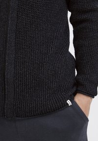 Svart stickad zip-sweater med texturerat mönster, sidofickor och en liten etikett vid fållen. Matchad med mörka byxor.