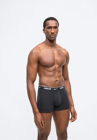 Μυώδης άνδρας όρθιος, φοράει μαύρα εσώρουχα μπόξερ Nike, κοιτάζοντας ευθεία μπροστά πάνω σε απλό ανοιχτό φόντο.