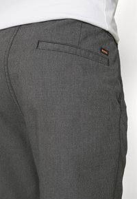 Pantalon gris à motifs avec une texture subtile, une poche arrière avec un accent noir et une petite étiquette logo orange.