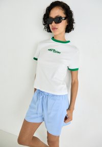T-shirt bianco a maniche corte con bordi verdi e logo "drive"; abbinato a pantaloni corti azzurri con coulisse. Modello che indossa grandi occhiali da sole neri.