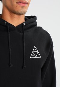 Czarna bluza z kapturem z mieszanki bawełny z regulowanymi sznurkami, z białym trójkątnym logo na przodzie, zawierającym litery 'H', 'U' i 'F'.