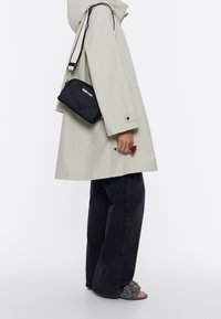 Lys grå vandtæt frakke, sort crossbody taske med logo, mørke jeans, grå blomstrede sandaler. Rød rosenring på hånden. Simpel, funktionel design.