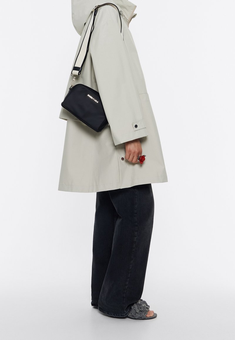 Lys grå vandtæt frakke, sort crossbody taske med logo, mørke jeans, grå blomstrede sandaler. Rød rosenring på hånden. Simpel, funktionel design.