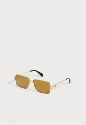 Lunettes de soleil rectangulaires à monture dorée avec verres bruns et embouts noirs sur des branches fines en métal, exposées sur une surface blanche.