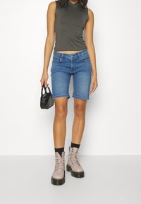 Mujer con un top corto sin mangas de color oliva oscuro, pantalones cortos de mezclilla azules hasta la rodilla, botas de encaje rosa pálido y sosteniendo un pequeño bolso negro.