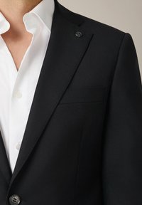 Czarny żakiet z wyraźnymi ramionami, zapięciem na jeden guzik i klapami. Posiada kieszeń na piersi oraz błyszczący przypinek na klapie. Pod spód założona biała koszula.