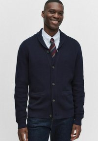 Marinblå cardigan med sjalkrage, ribbad textur, framknappar och två fickor, bärs över en skjorta med krage och slips, ihop med mörka jeans.