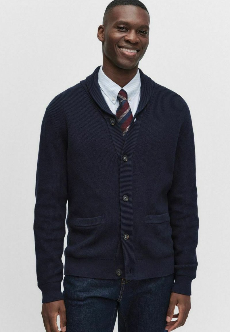 Marinblå cardigan med sjalkrage, ribbad textur, framknappar och två fickor, bärs över en skjorta med krage och slips, ihop med mörka jeans.