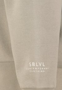 Lichtgrijze sweatshirt met ribgebreide manchetten en zoom. Voorzien van een wit label met de tekst "SBLVL CONTEMPORARY CLOTHING" op de onderarm. Zachte textuur.
