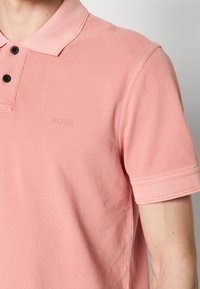 Gros plan d'une personne portant un polo rose clair avec deux boutons noirs et un logo "BOSS" subtil sur la poitrine.